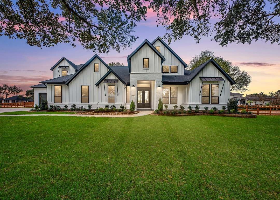 Modern white farmhouse with prominent front-facing gables, a broad green lawn, and mature trees at sunset